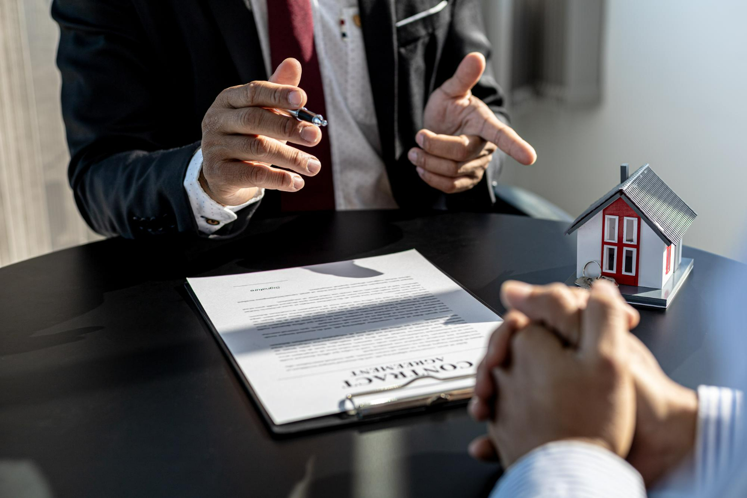 Hands gesturing during a real estate negotiation over a contract, with a miniature house model and a signed agreement on the table, reflecting legal discussions in Carlsbad.