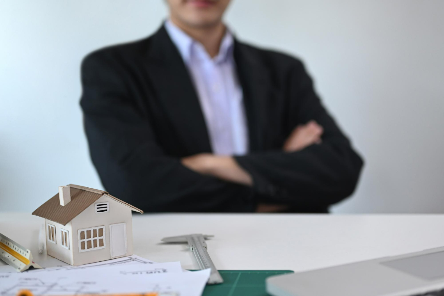 Lawyer in a suit with crossed arms, blurred background, model house and real estate documents on the table, symbolizing real estate law and property transactions in Carlsbad.