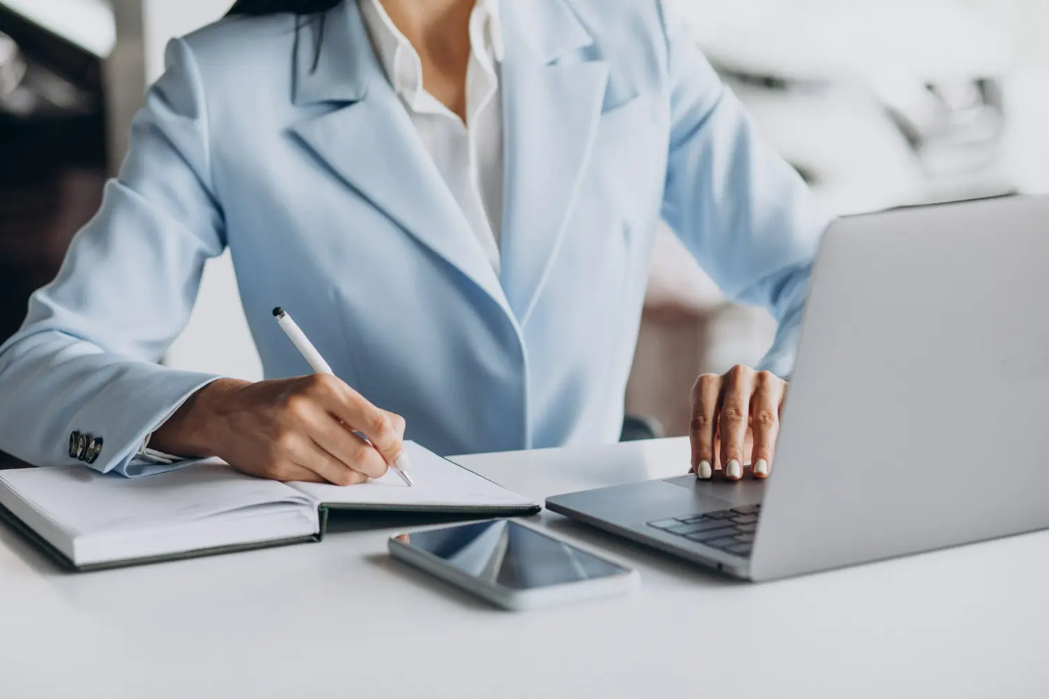 A businesswoman in a light blue suit writing notes in a notebook while working on a laptop at her desk