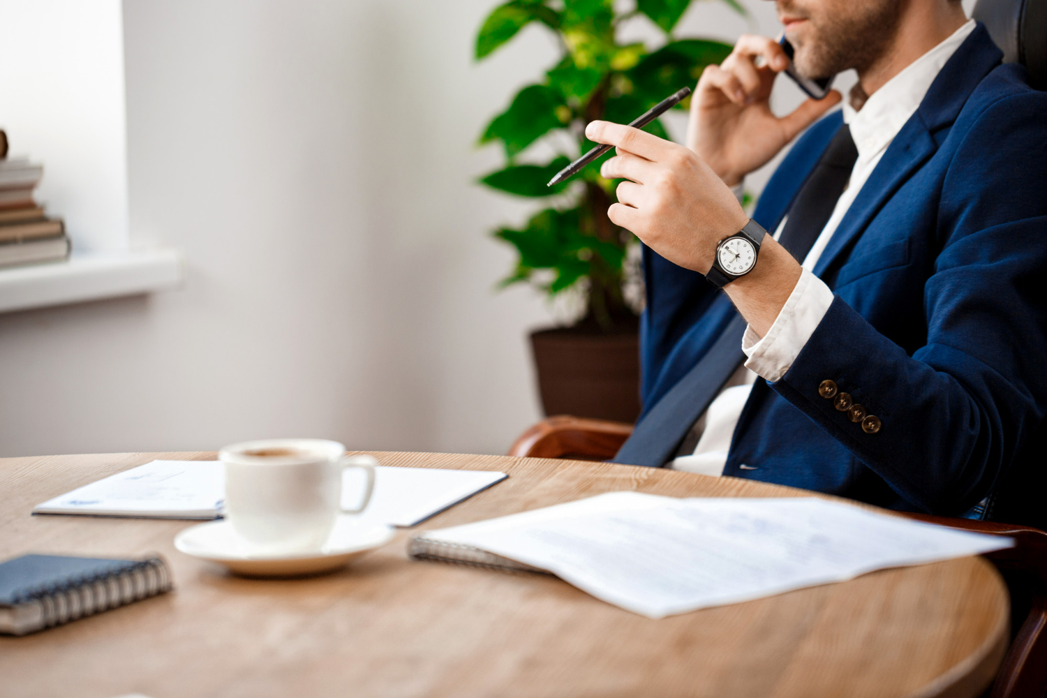 Business professional on phone call, holding pen, surrounded by legal documents and coffee cup, in office setting with plants, emphasizing legal services and contract discussions.