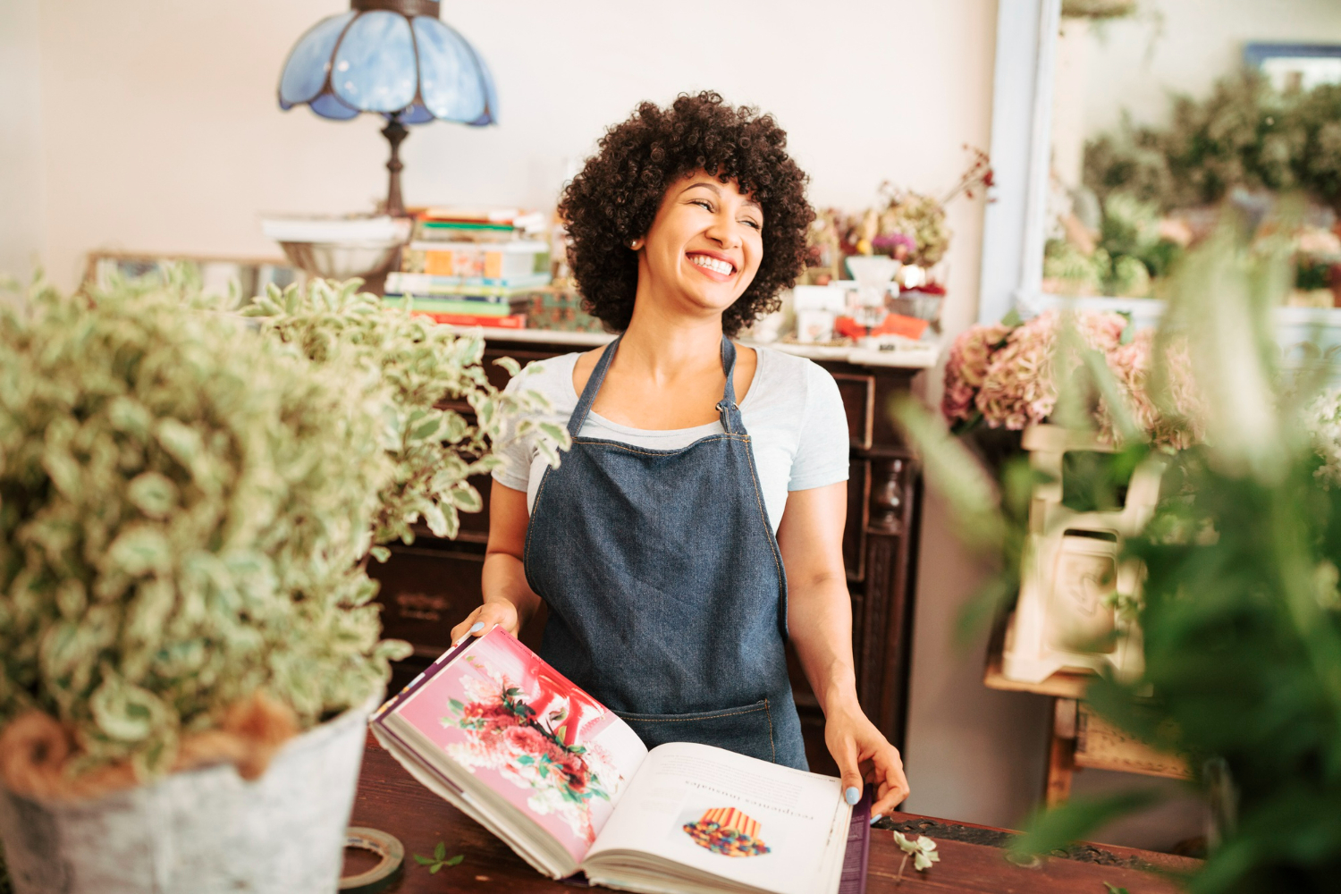 Smiling woman in an apron holding an open book, surrounded by plants in a cozy workspace, illustrating the importance of expert advice in real estate negotiations and lease agreements.