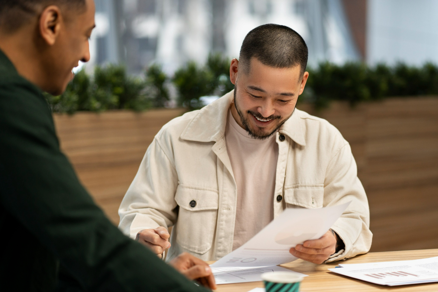 Two men engaged in a discussion at a table, one smiling while reviewing documents, emphasizing collaboration and business planning in a modern office setting.