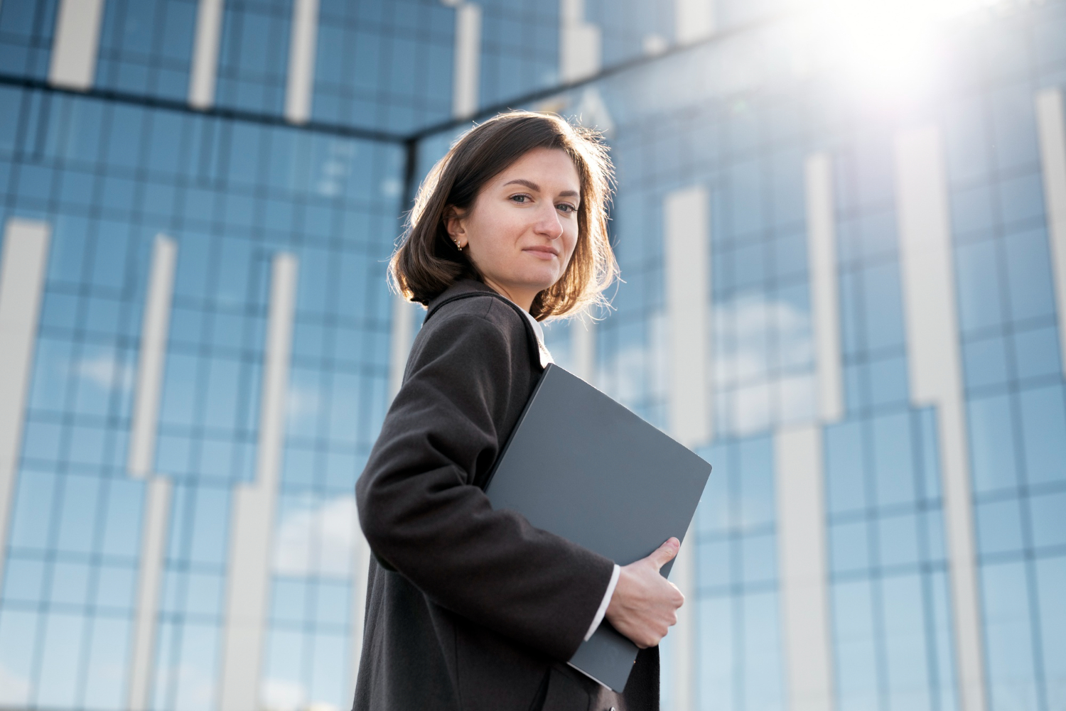 A business professional in a dark coat holding a grey folder, standing confidently in front of a modern glass office building with sunlight reflecting off the windows.