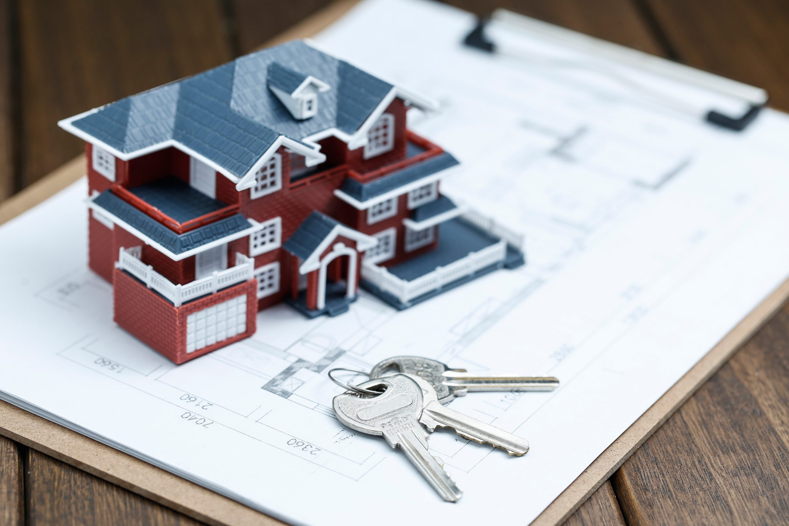 A small model of a red and white house sits on a clipboard with architectural blueprints and a set of silver house keys, representing the real estate planning and purchase process under California Prop 13
