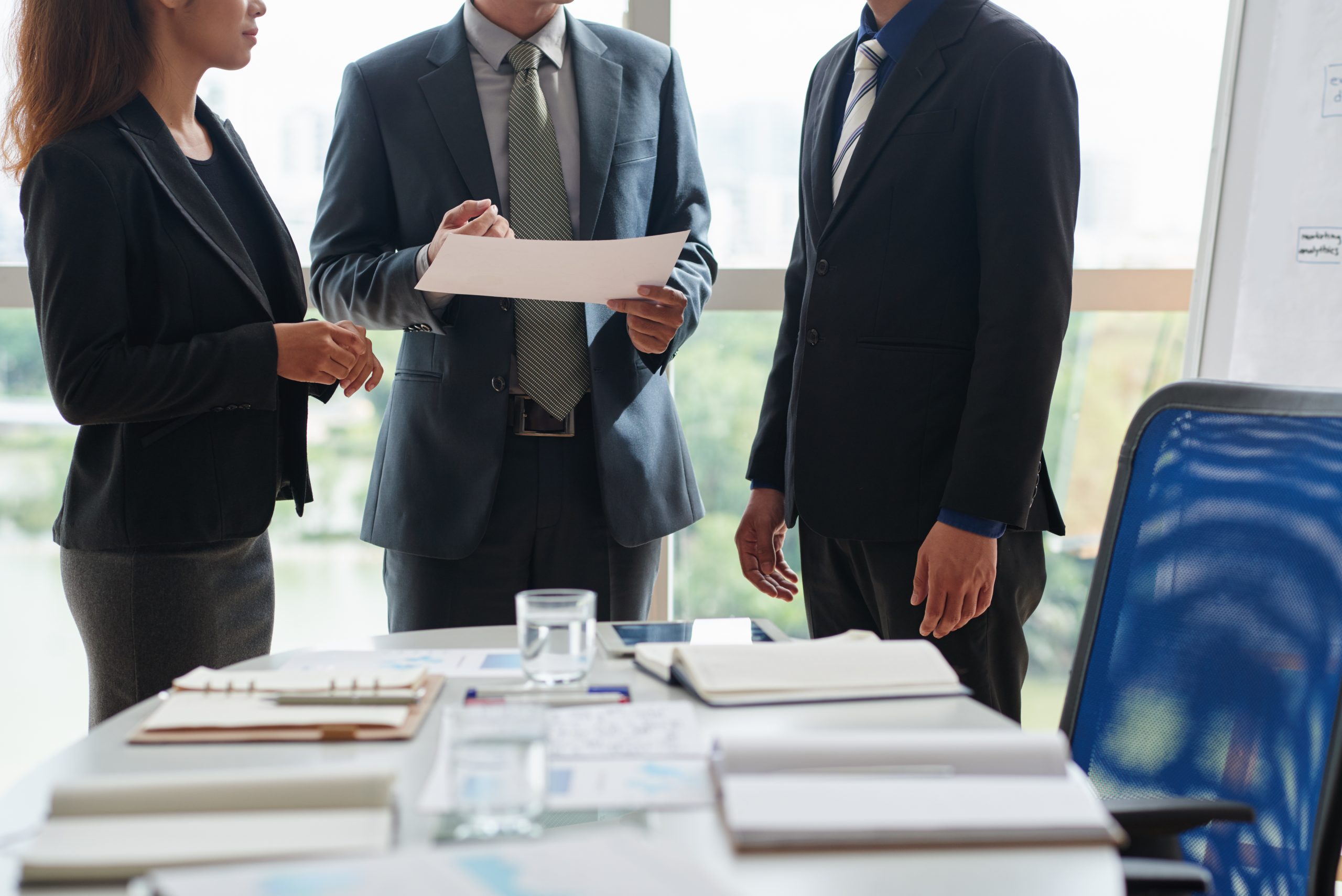 Three professionals in business attire stand around a bright office table, reviewing a document together to illustrate a joint venture where multiple parties pool resources and expertise to achieve a common real estate goal