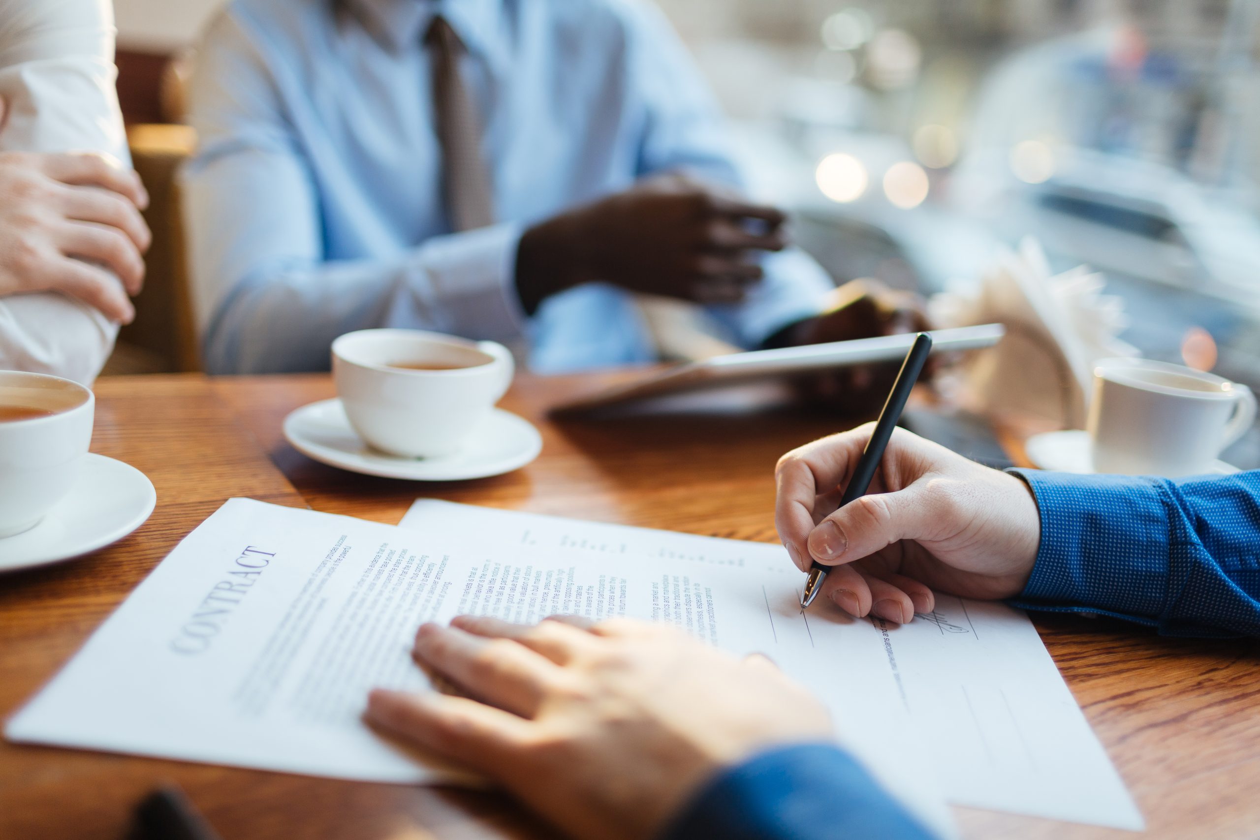 A close-up shot of a person's hand signing a document clearly labeled "CONTRACT" on a wooden table. In the background, other professionals are blurred, seated with coffee cups and a tablet, representing the finalization of a business agreement and the importance of clear, non-ambiguous contract language