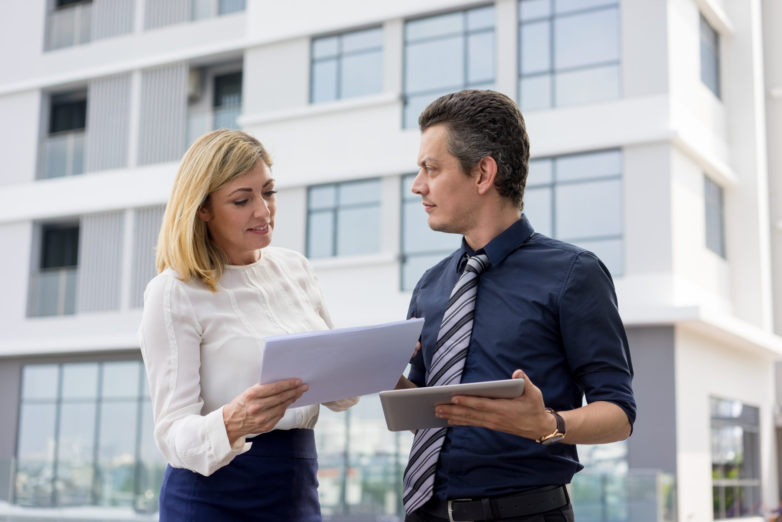 Two professionals stand outside a commercial building reviewing documents, discussing details related to due diligence in commercial real estate transactions