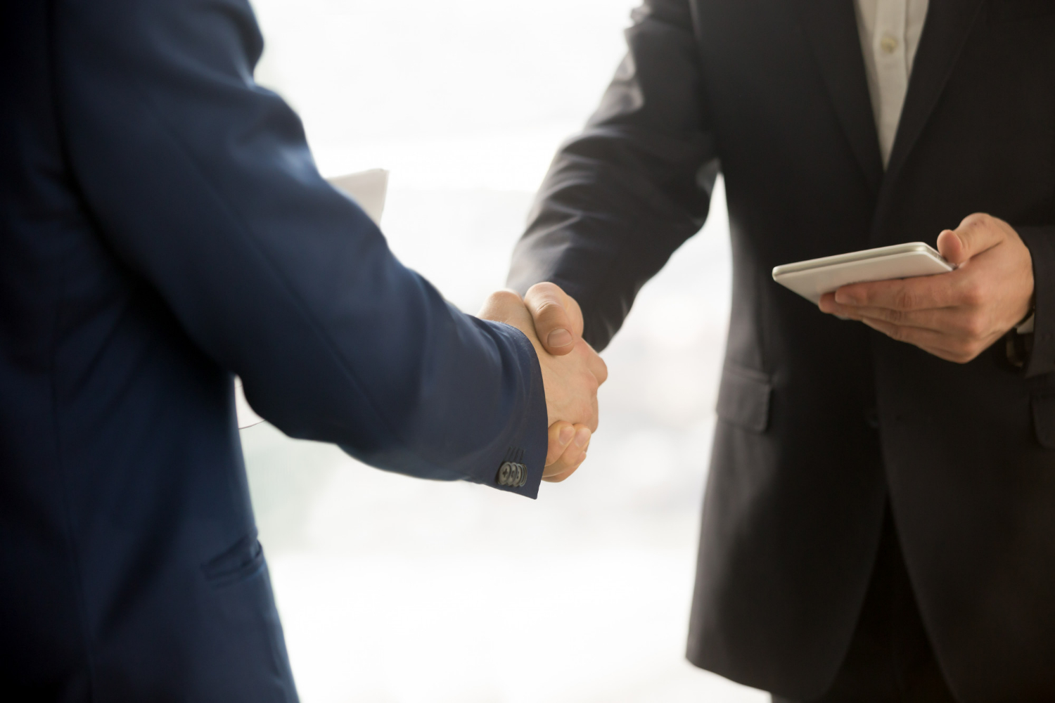 A close-up of a handshake between professionals while another reviews a tablet, representing formal agreements and the importance of keeping partnership terms current